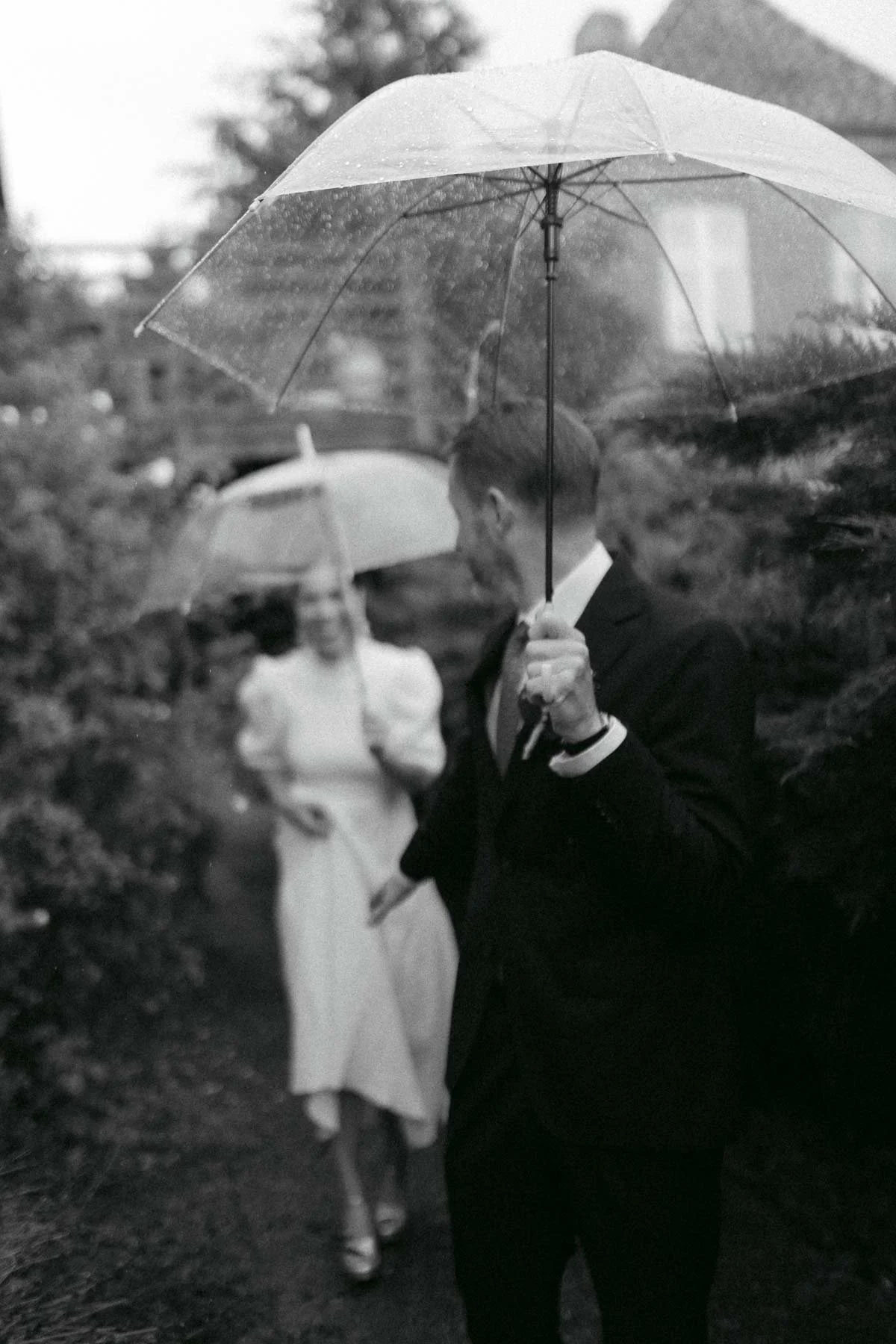 A man in a suit holding an umbrella with a woman in a dress in the background, both standing outdoors in the rain, with blurred trees and buildings behind them.