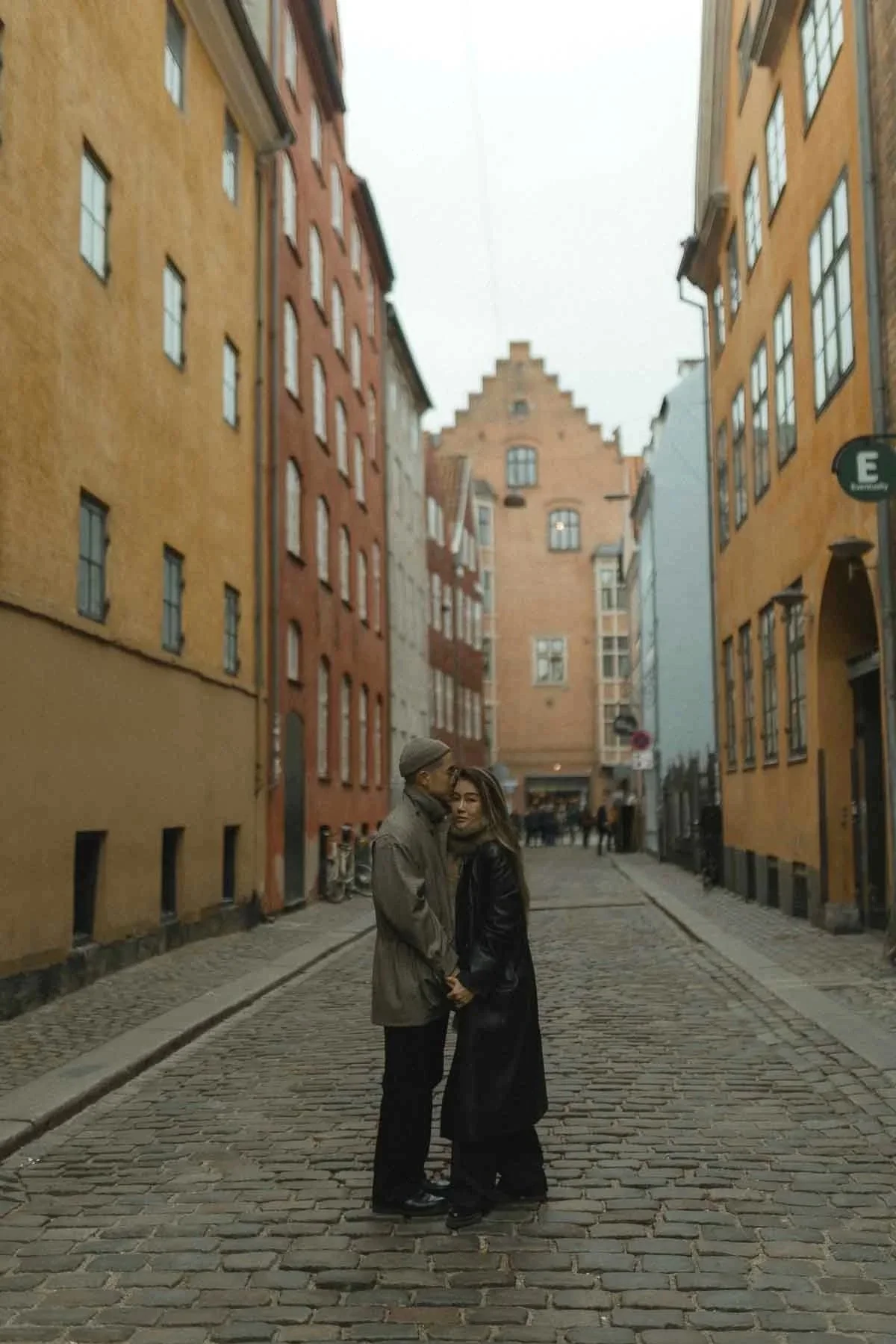 A couple stands close together on a cobblestone street lined with colorful, historic European-style buildings, with a tall, stepped gable building in the background, during daytime.