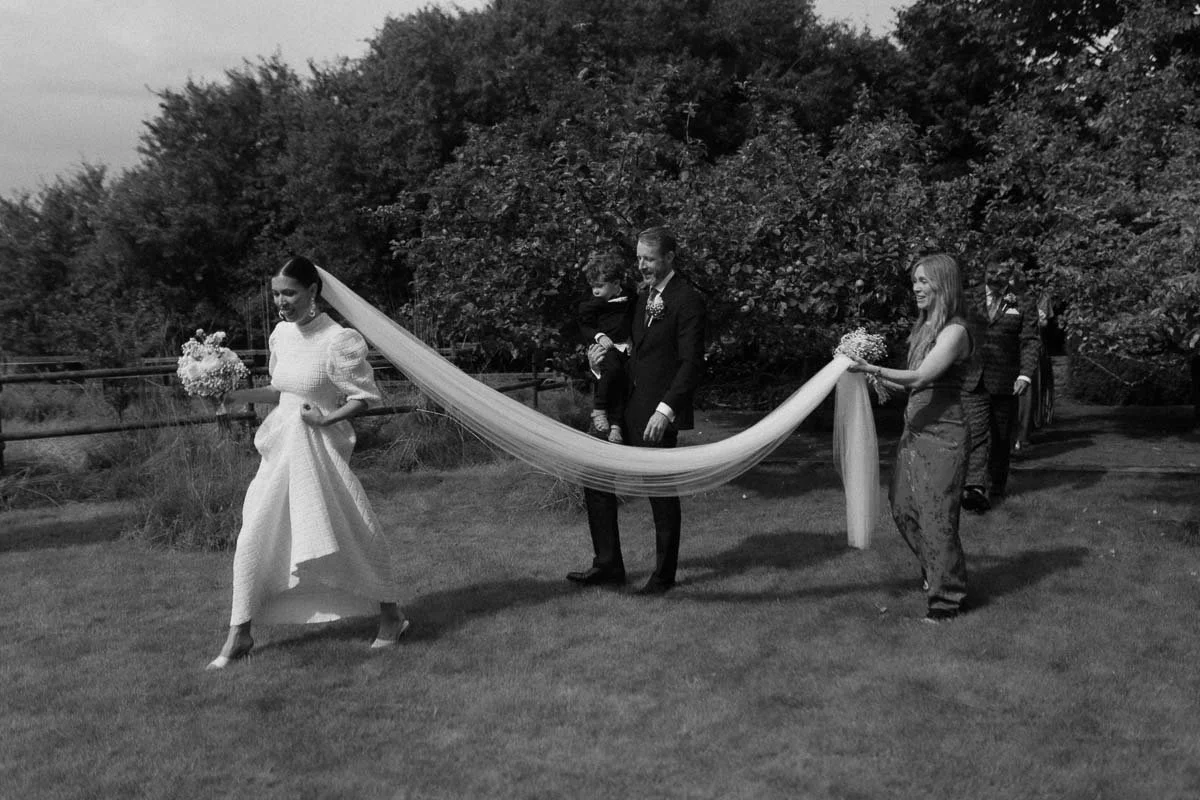 Copenhagen Wedding Photographer. Black and white photo of a wedding procession outdoors with a bride in a white dress holding a bouquet, a man in a suit holding a young child, and a young woman in a dress holding flowers, walking on grass with trees 