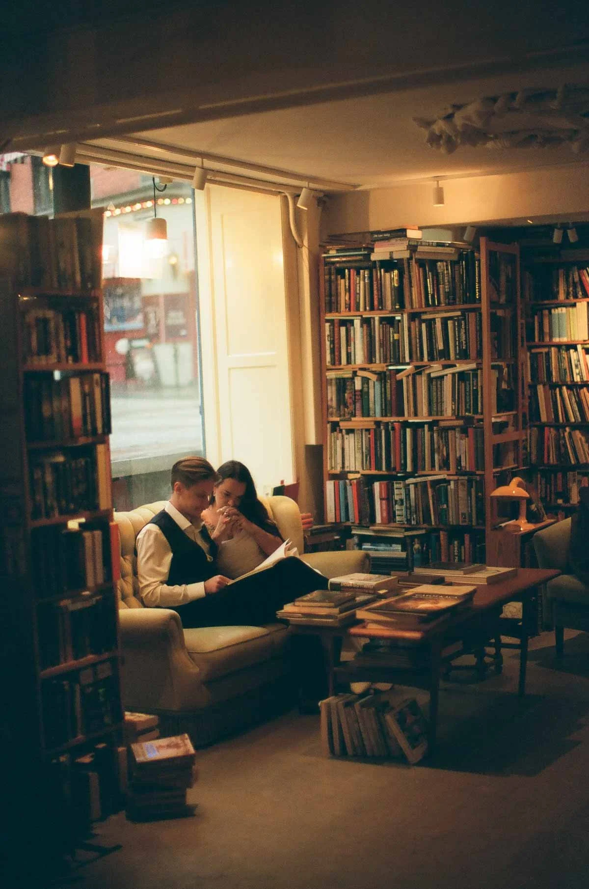 Copenhagen Wedding Photographer. Two people sitting on a couch in a cozy bookstore or library, reading and sharing a moment together, surrounded by bookshelves filled with books.