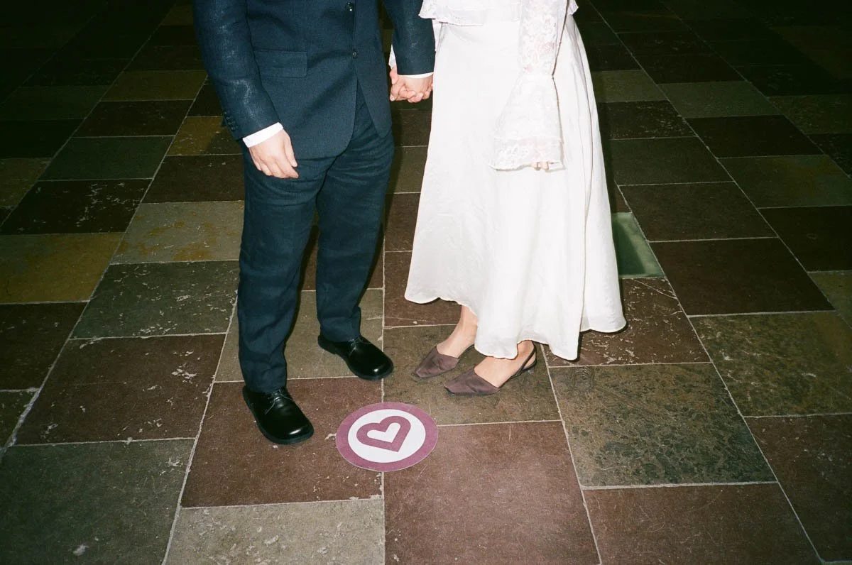 A close-up of a wedding couple holding hands, with only their lower bodies visible, standing on a tiled floor with a love heart symbol on the ground.