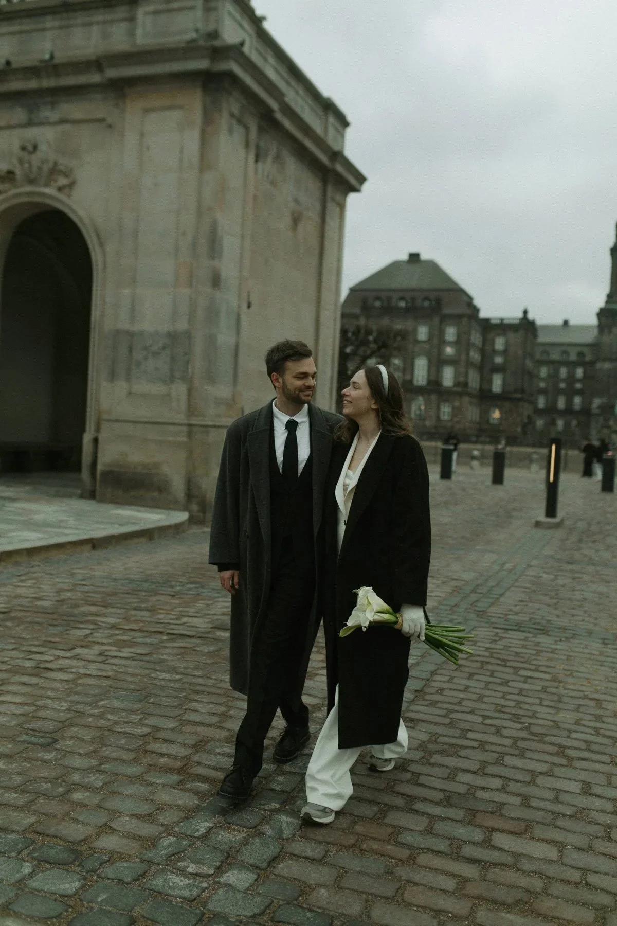 Copenhagen Wedding Photographer. A happy couple dressed in wedding attire walking on cobblestone street, woman holding white calla lilies, in an urban area with historic buildings.