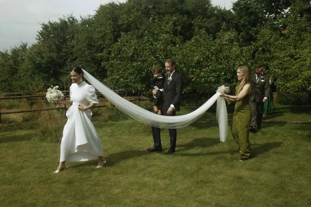 Bride in white dress holding a bouquet, walking outdoors on grass, with people and trees in the background, during a wedding celebration.