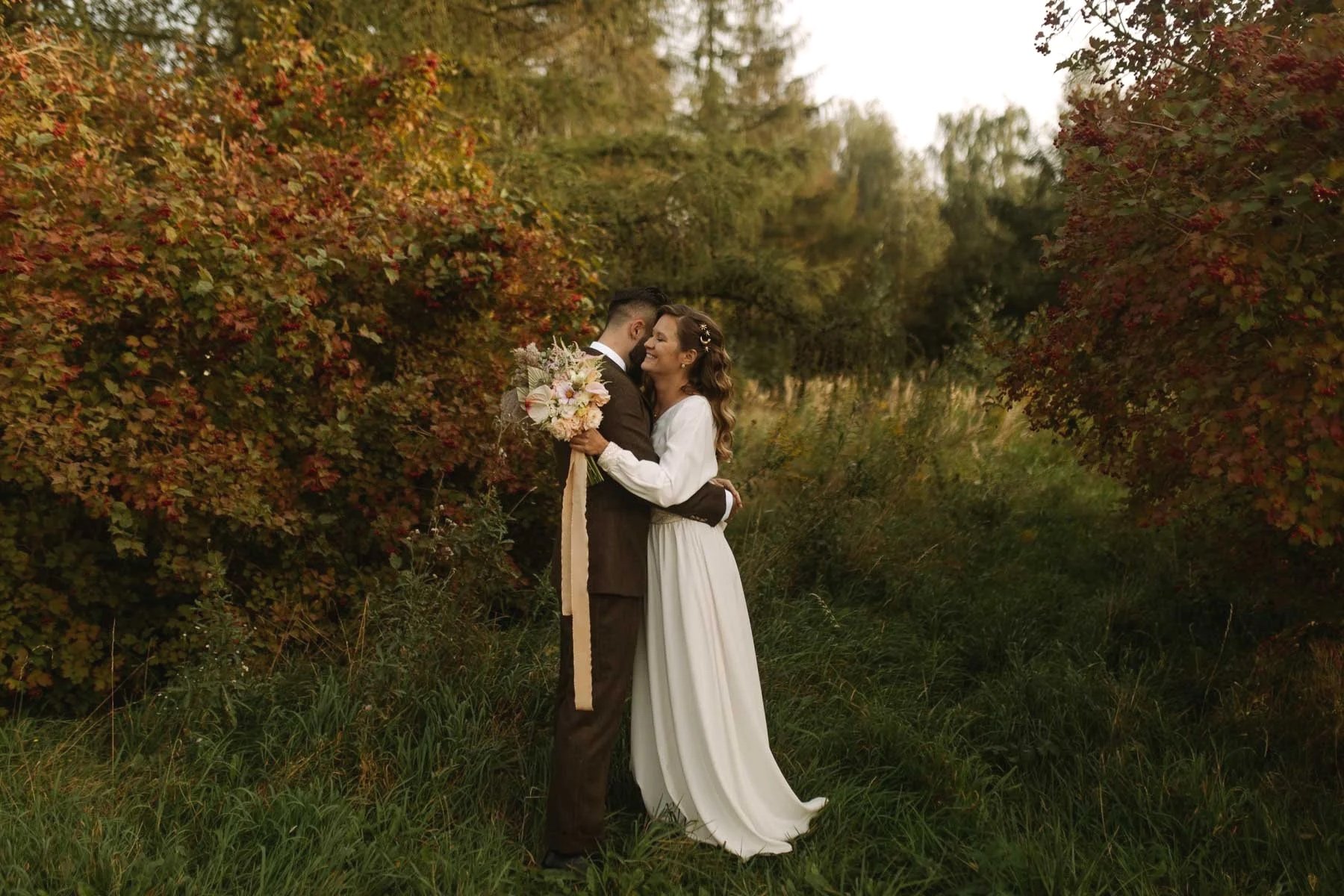 A newlywed couple embracing outdoors during fall, with colorful foliage in the background. The bride is holding a bouquet of flowers, and they are smiling at each other.