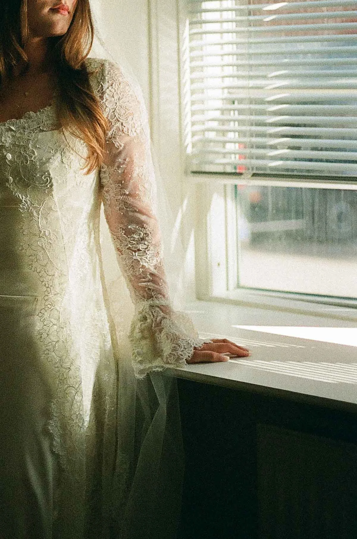 Copenhagen Wedding Photographer. A woman in a lace dress standing by a window with blinds, looking outside.
