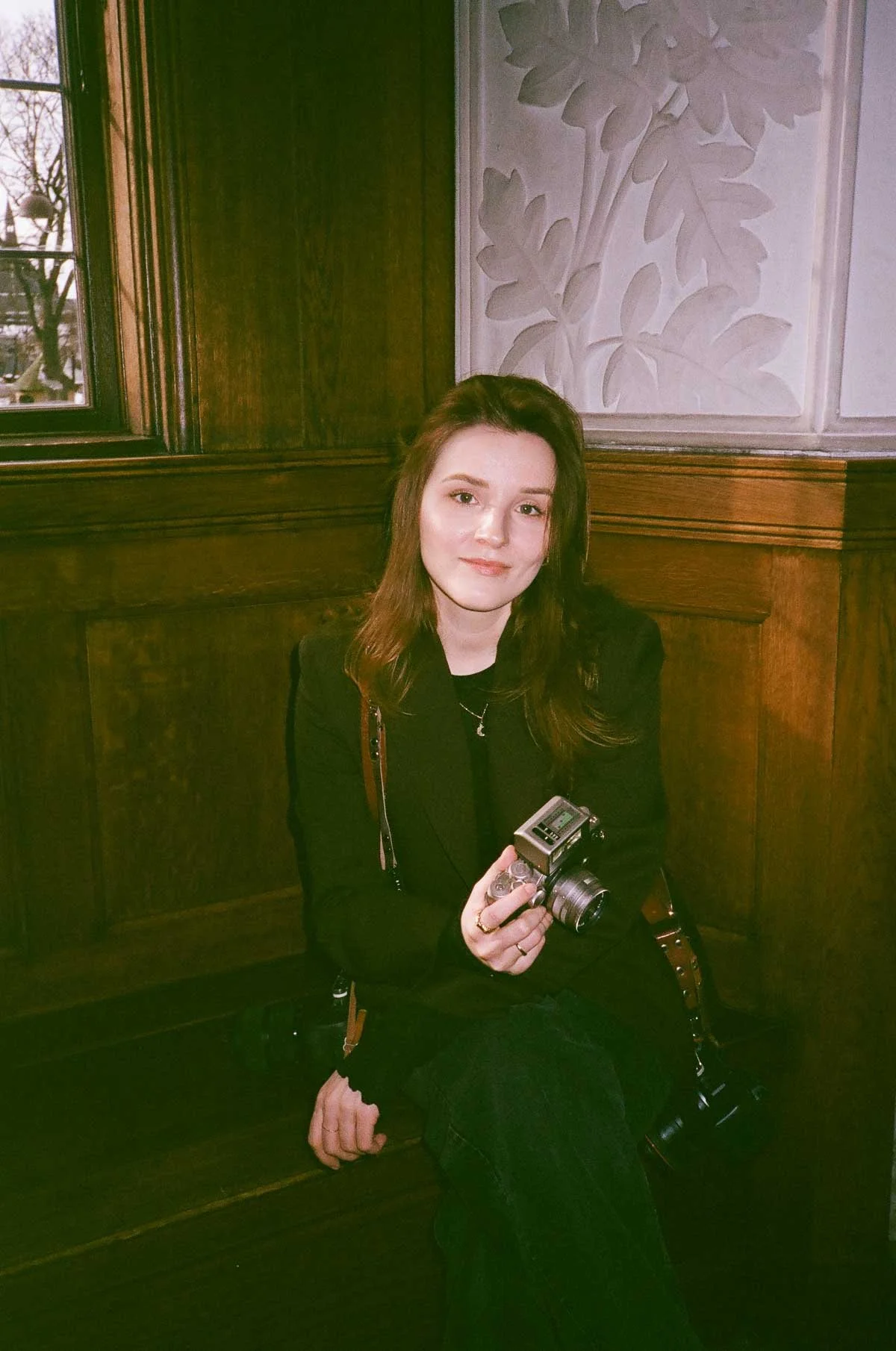 A young woman with light brown hair sitting on a wooden bench, holding a vintage film camera, in a room with wood paneled walls and decorative wall art.