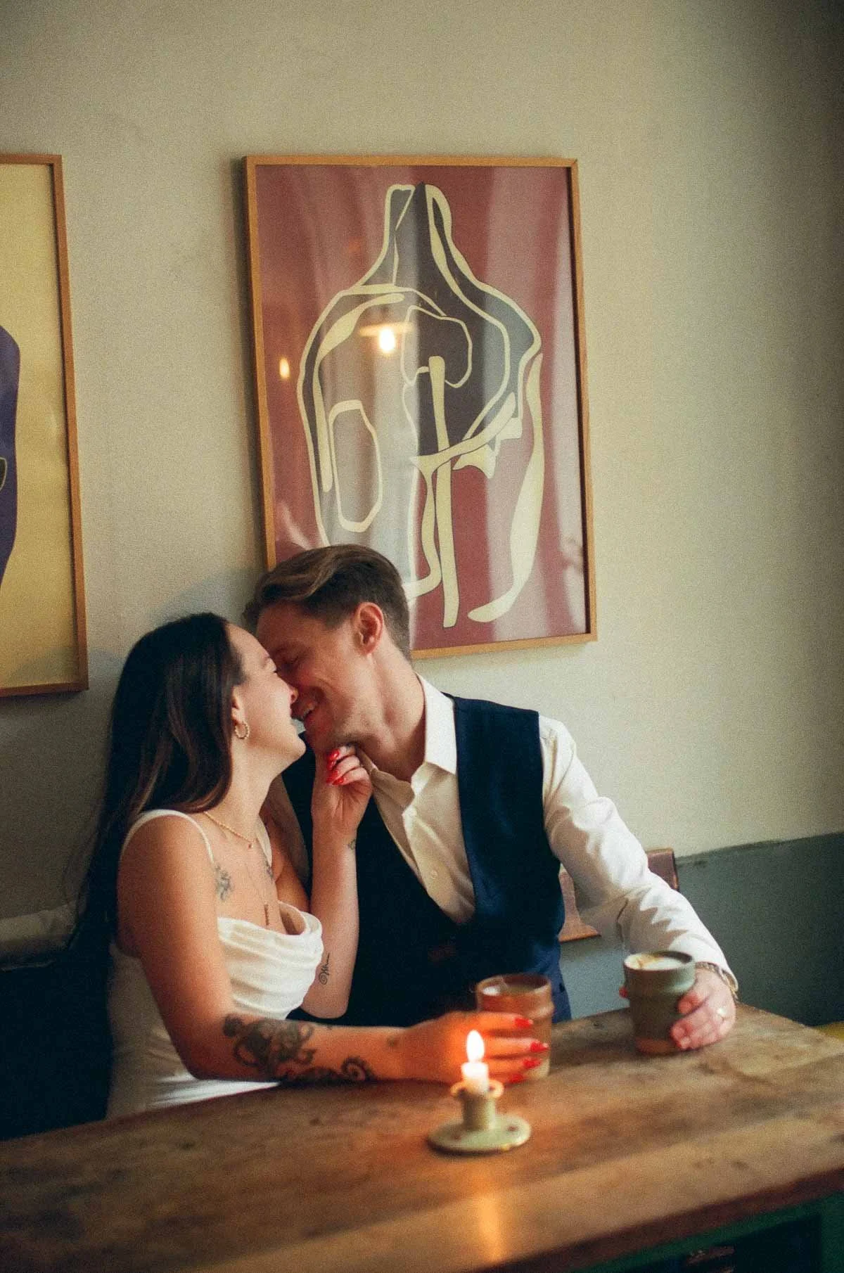Copenhagen Wedding Photographer. A couple sitting at a wooden table, smiling and close together, touching noses with a small candle on the table. The woman has tattoos and long dark hair, and the man has short hair and is wearing a white shirt with a