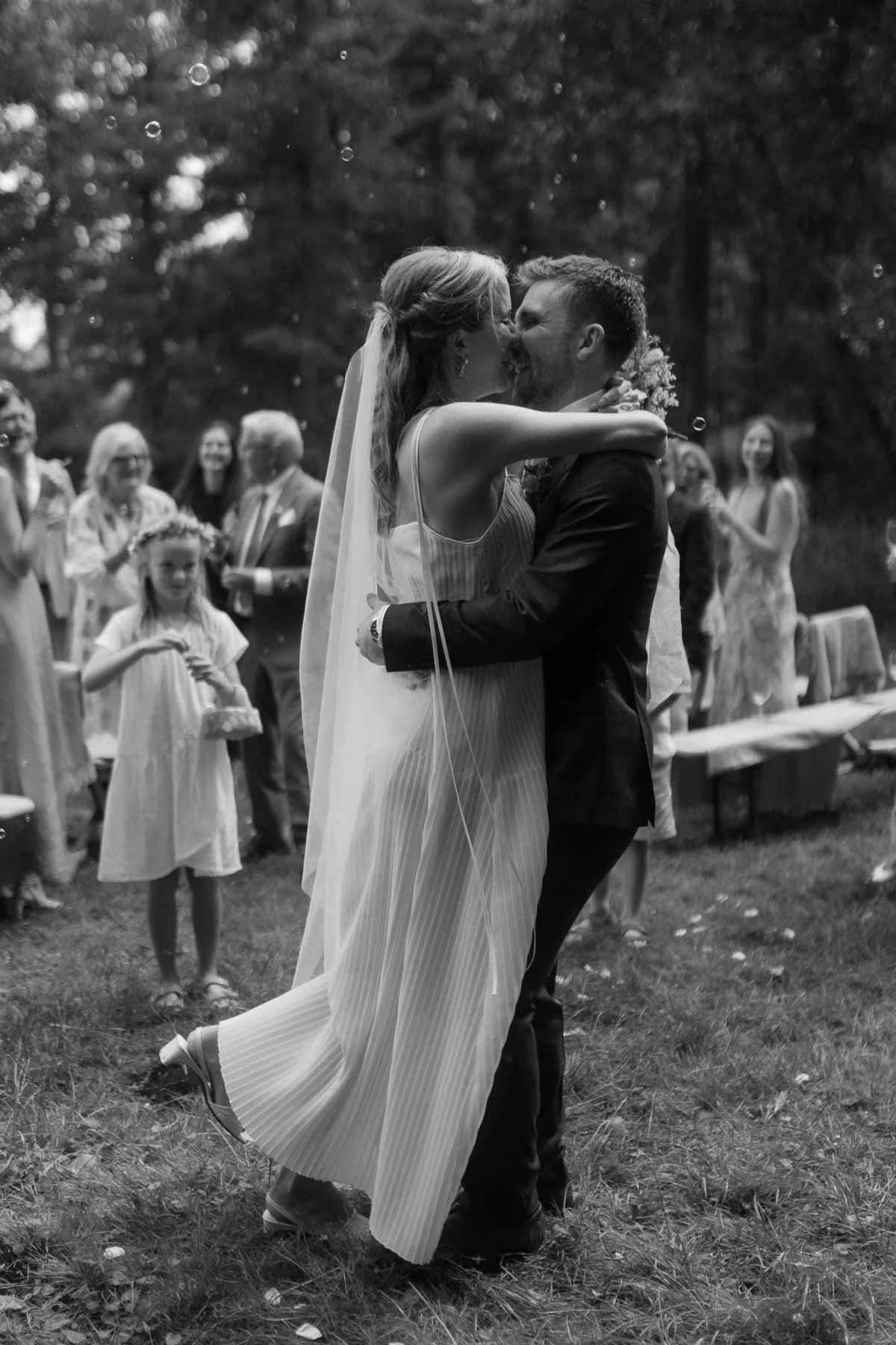 Copenhagen Wedding Photographer. A black-and-white photo of a bride and groom embracing and kissing during their wedding ceremony outdoors, surrounded by guests and trees.