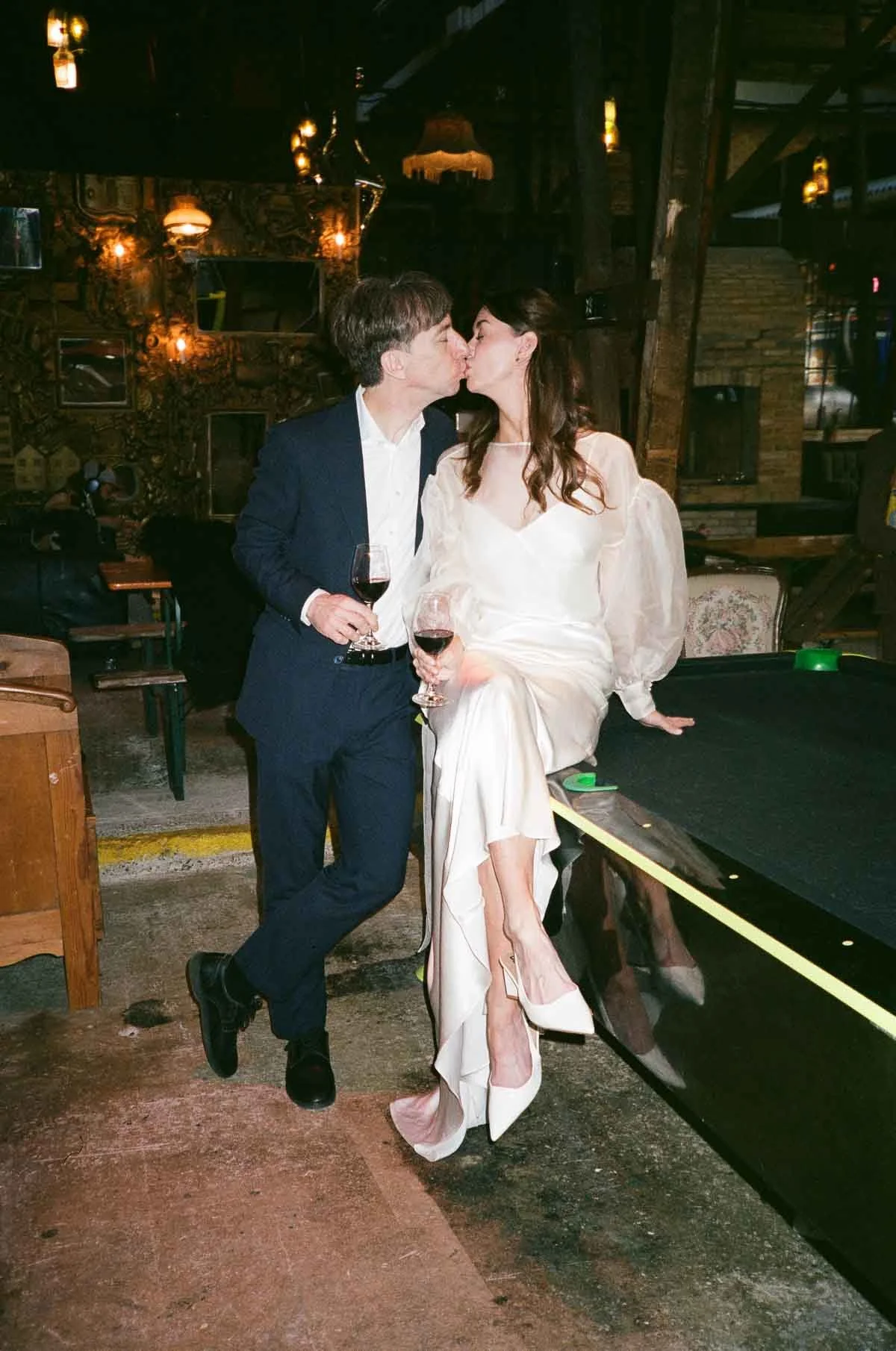 A man and woman in formal attire sharing a kiss while holding glasses of red wine in a dimly lit bar or restaurant with a rustic interior.