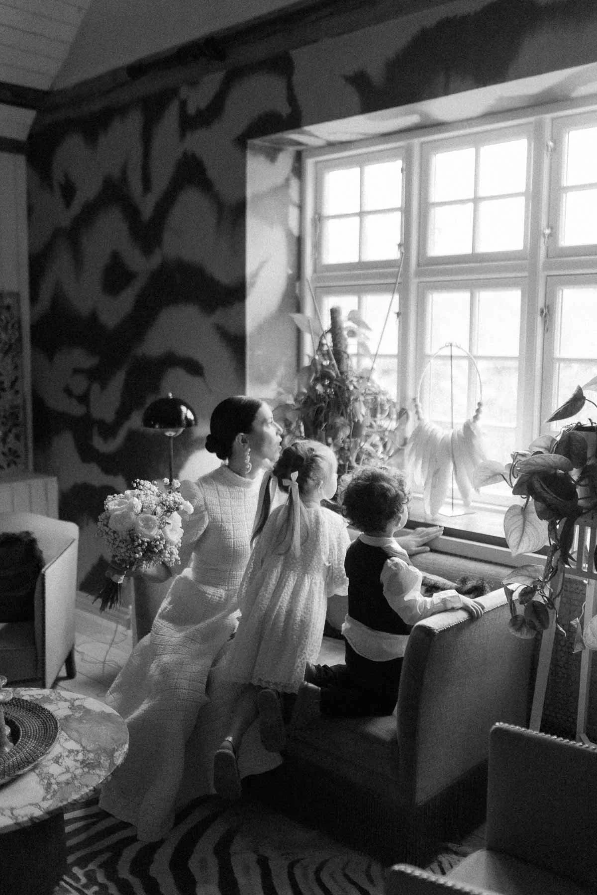 ACopenhagen Wedding Photographer.  woman and two children kneeling by a window, observing a cat statue with a hoop. The room has decorative plants, a patterned wall, and various furniture, with natural light coming through the window.