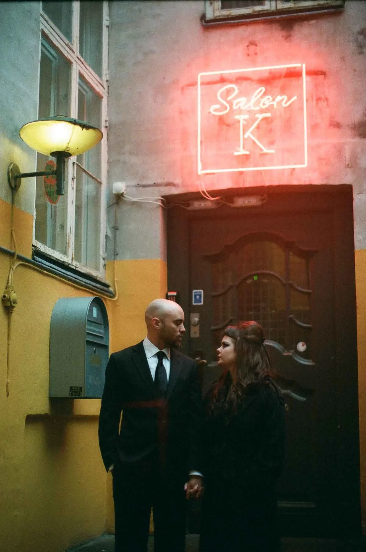 Copenhagen Wedding Photographer. A man in a black suit and a woman in black clothing standing close together in an alley, holding hands. The alley has a yellow lower wall, a gray upper wall, a window with a lamp, and a neon sign that reads 'Salon K' 