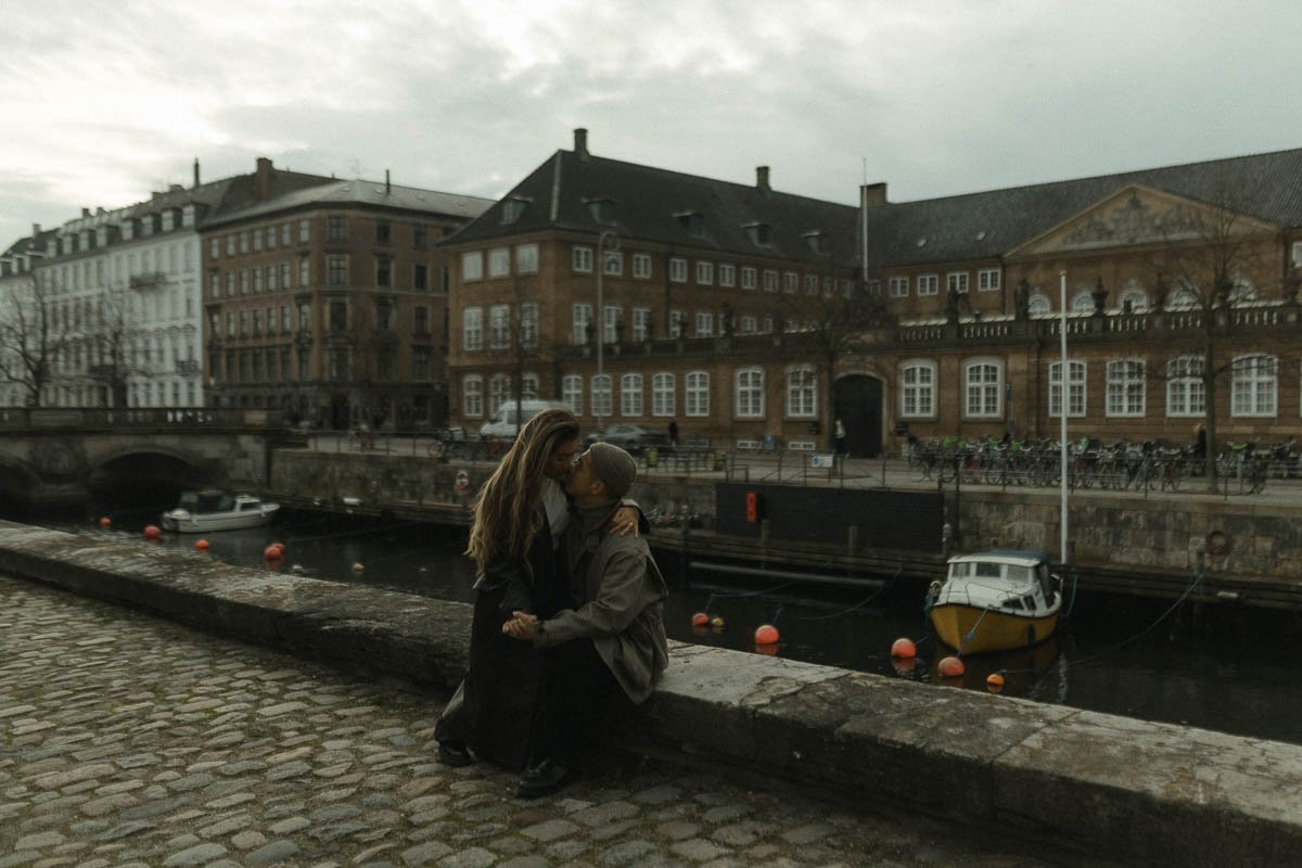 A man and woman share an intimate moment by a canal, with historic brick buildings and a small boat in the background in a city setting.