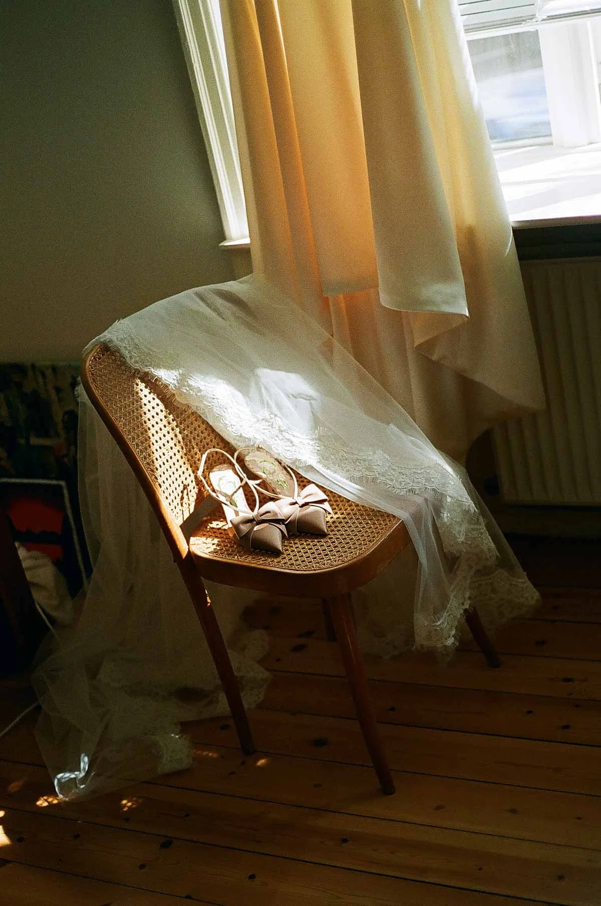 Copenhagen Wedding Photographer. A pair of white wedding shoes with bows placed on a wooden chair draped with a lace veil, set in front of a window with sunlight streaming through cream-colored curtains.