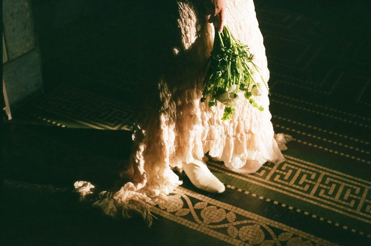 Person in a long, white, lace dress holding a bouquet of white flowers, standing on a decorative tiled floor in dim lighting.