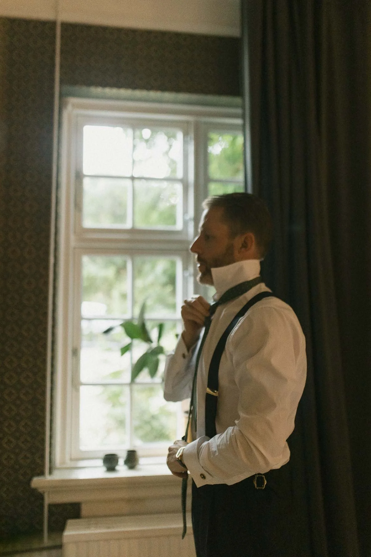 A man in formal attire, with a bow tie and suspenders, adjusting his collar while standing indoors near a window with greenery outside.