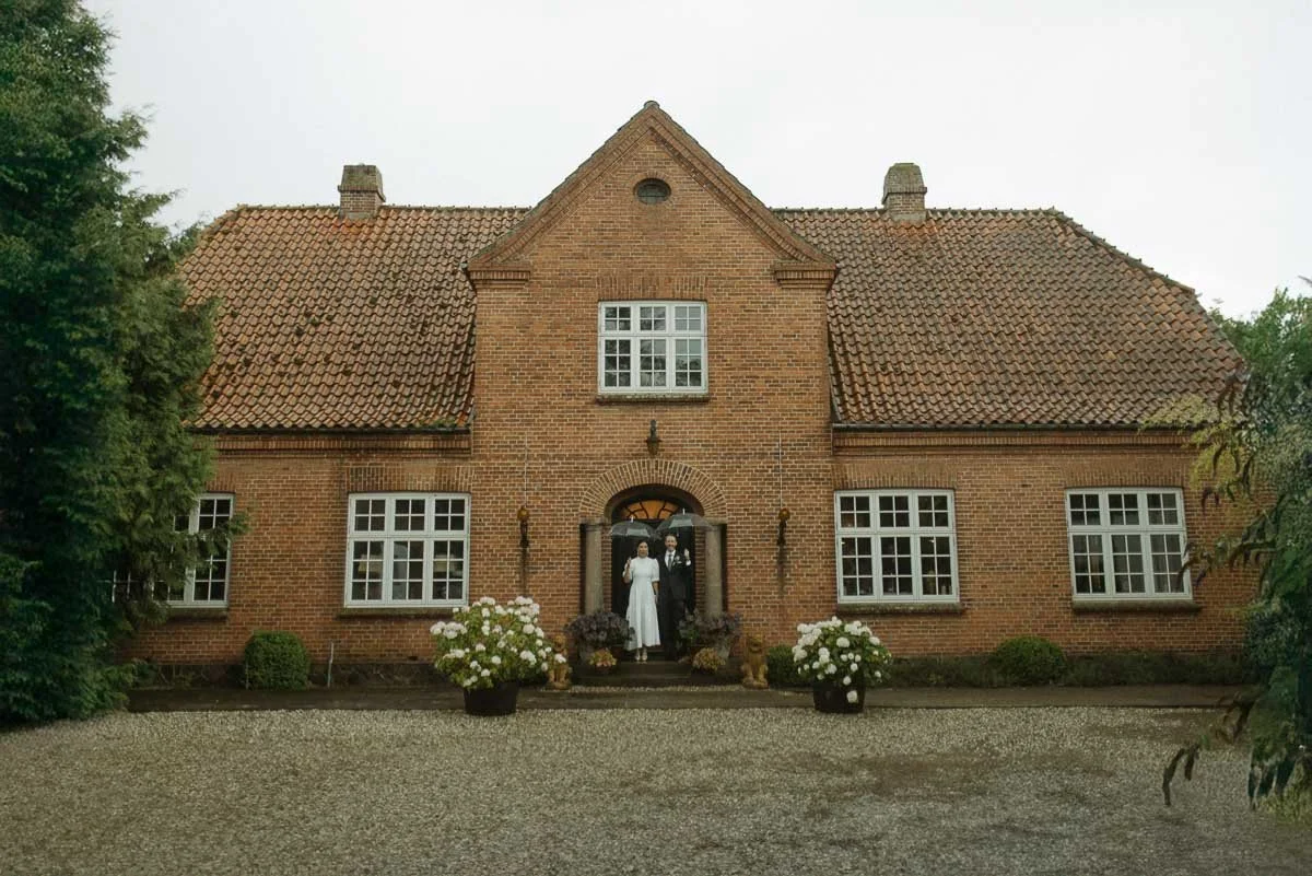 Bride and groom standing at the entrance of a brick house with white-framed windows and a gravel driveway.
