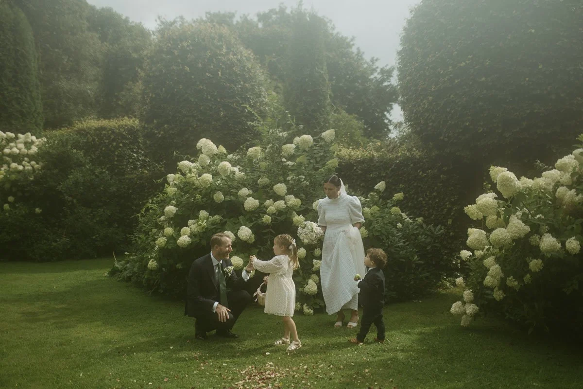 Copenhagen Wedding Photographer. A couple dressed in wedding attire with two young children in a garden with white flowers and greenery.