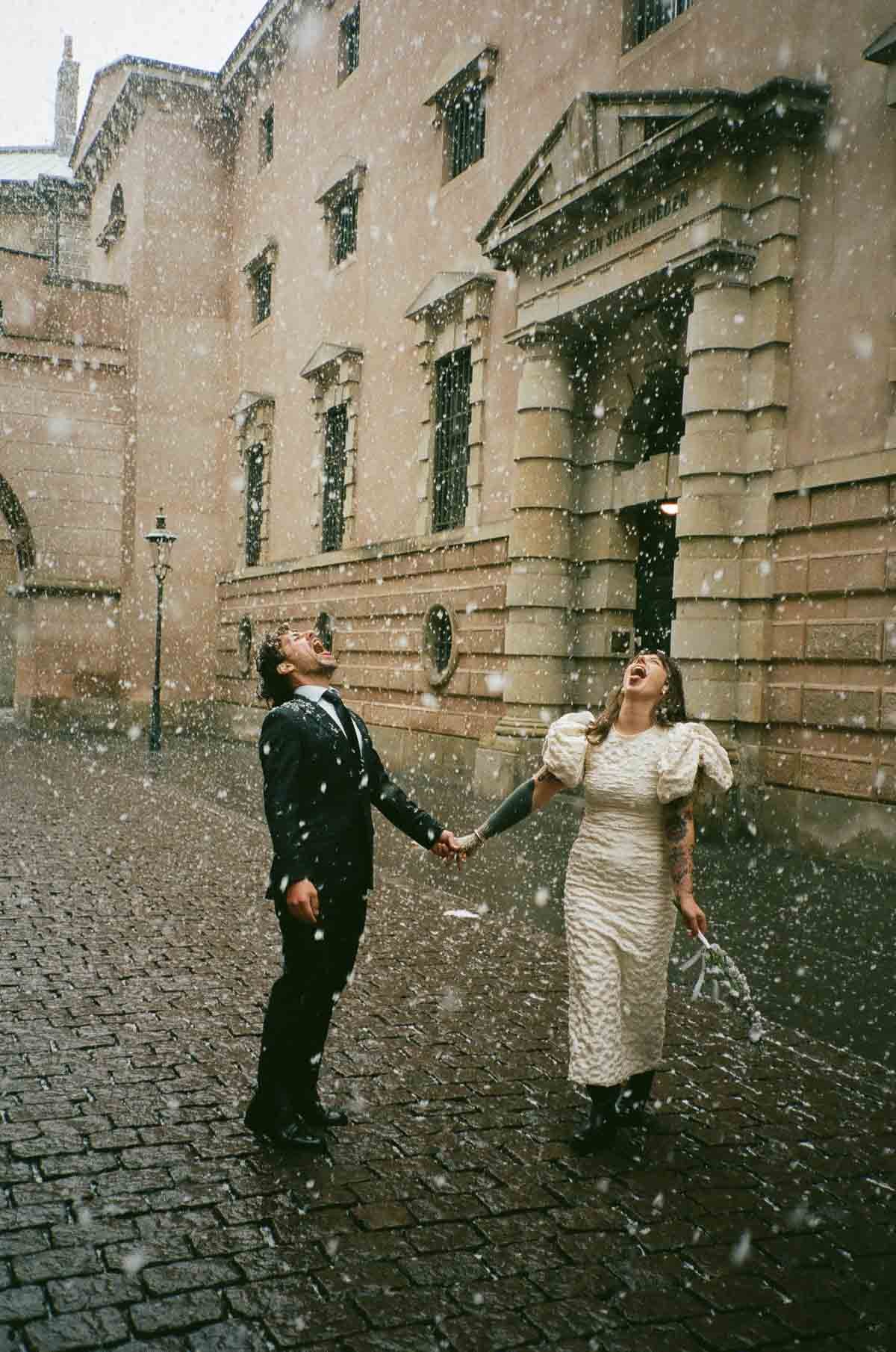 Copenhagen Wedding Photographer.  A joyful couple in wedding attire holding hands and laughing in a rainstorm on a cobblestone street in front of a historic building.