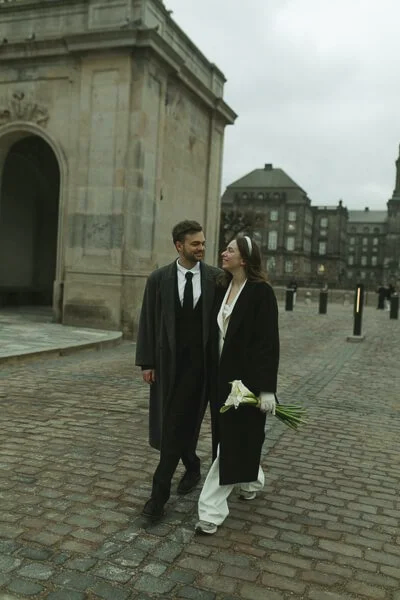 A couple dressed in black and white outfits walking on cobblestone in front of a historic building.