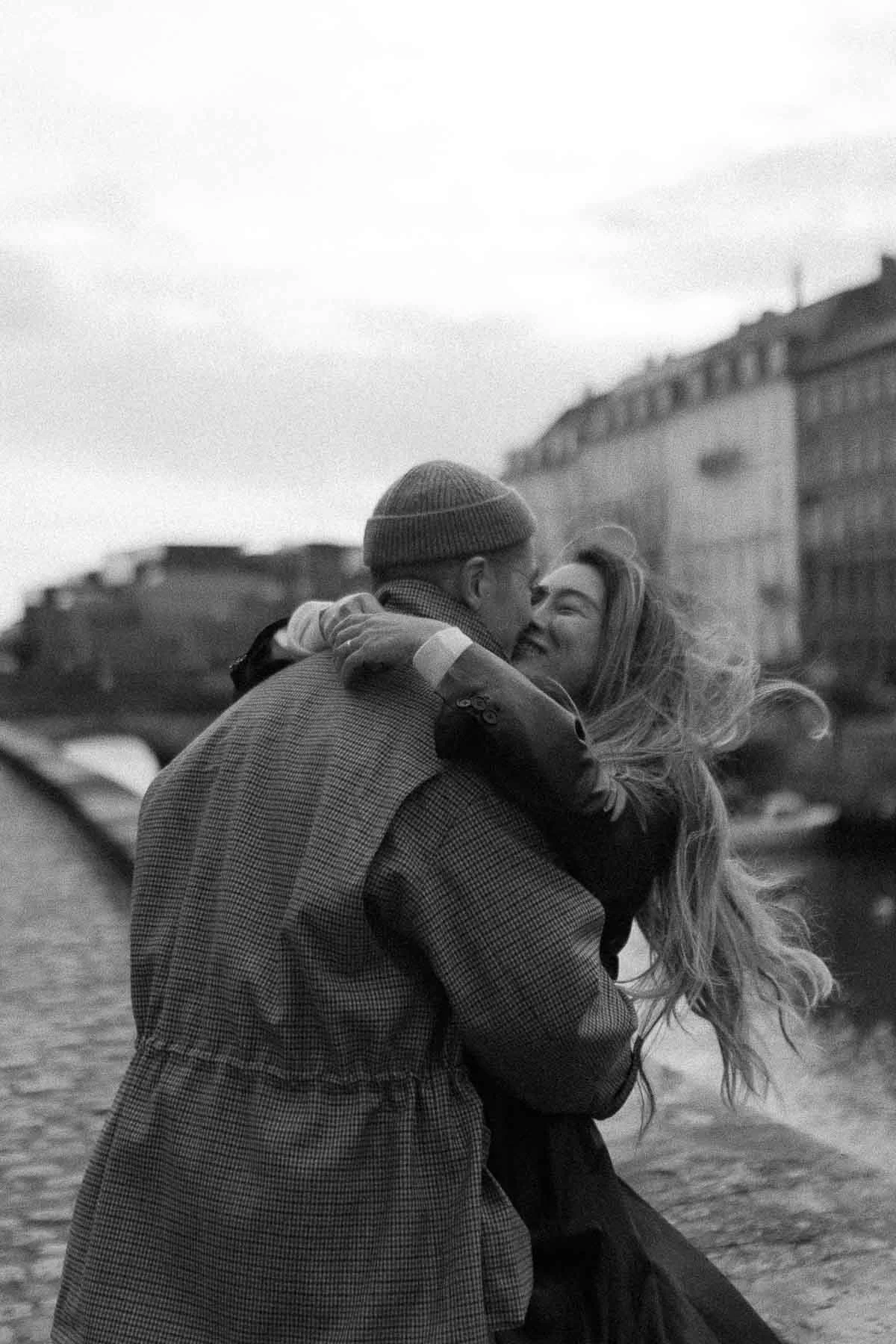 A black and white photo of a man and woman embracing by the water, with city buildings in the background.