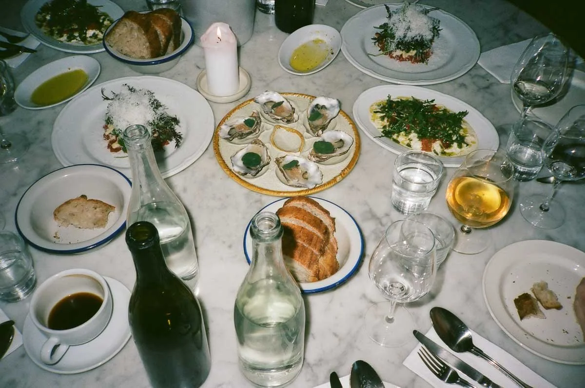 A table set with various dishes including oysters, bread, salad, and sauces. There are also several glasses of wine and water, a lit candle, and utensils.