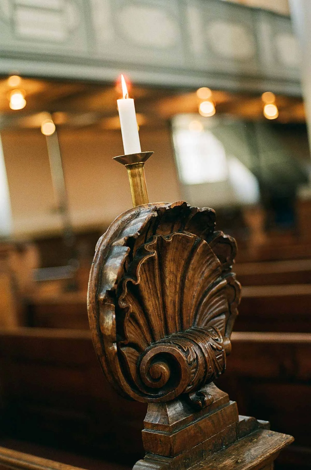 Copenhagen Wedding Photographer. A lit white candle on a brass candlestick holder, placed on a carved wooden pew in a church.