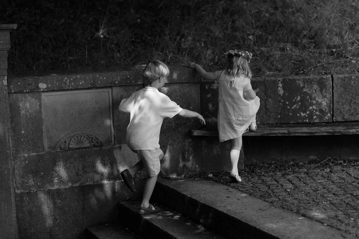Copenhagen Wedding Photographer.  Two young children, a boy and a girl, exploring near a stone wall with a wooded area in the background, at night. The girl is wearing a dress and a floral crown, and the boy is wearing a short-sleeved shirt and short