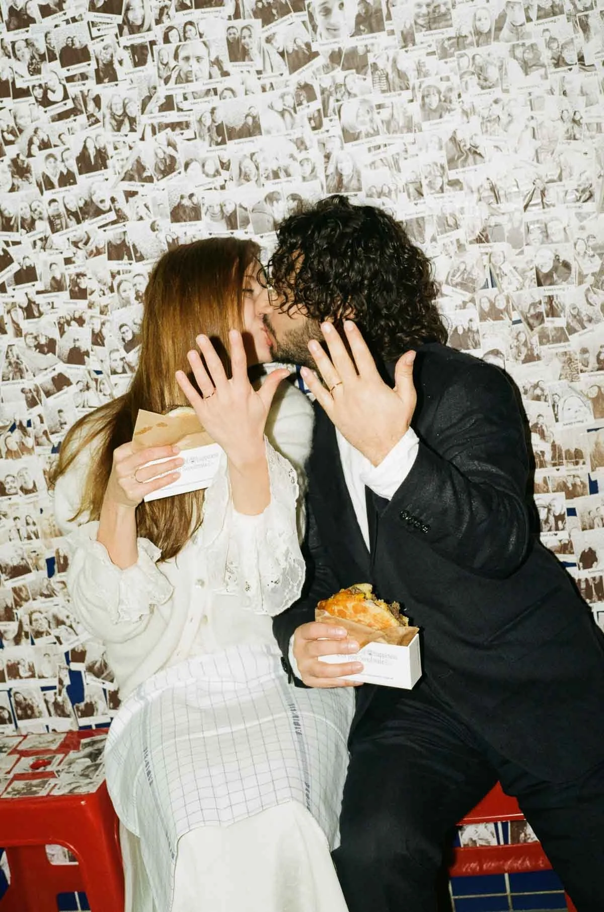 A couple is sharing a romantic moment, sitting close to each other while holding food, with a wall covered in black and white photos in the background.