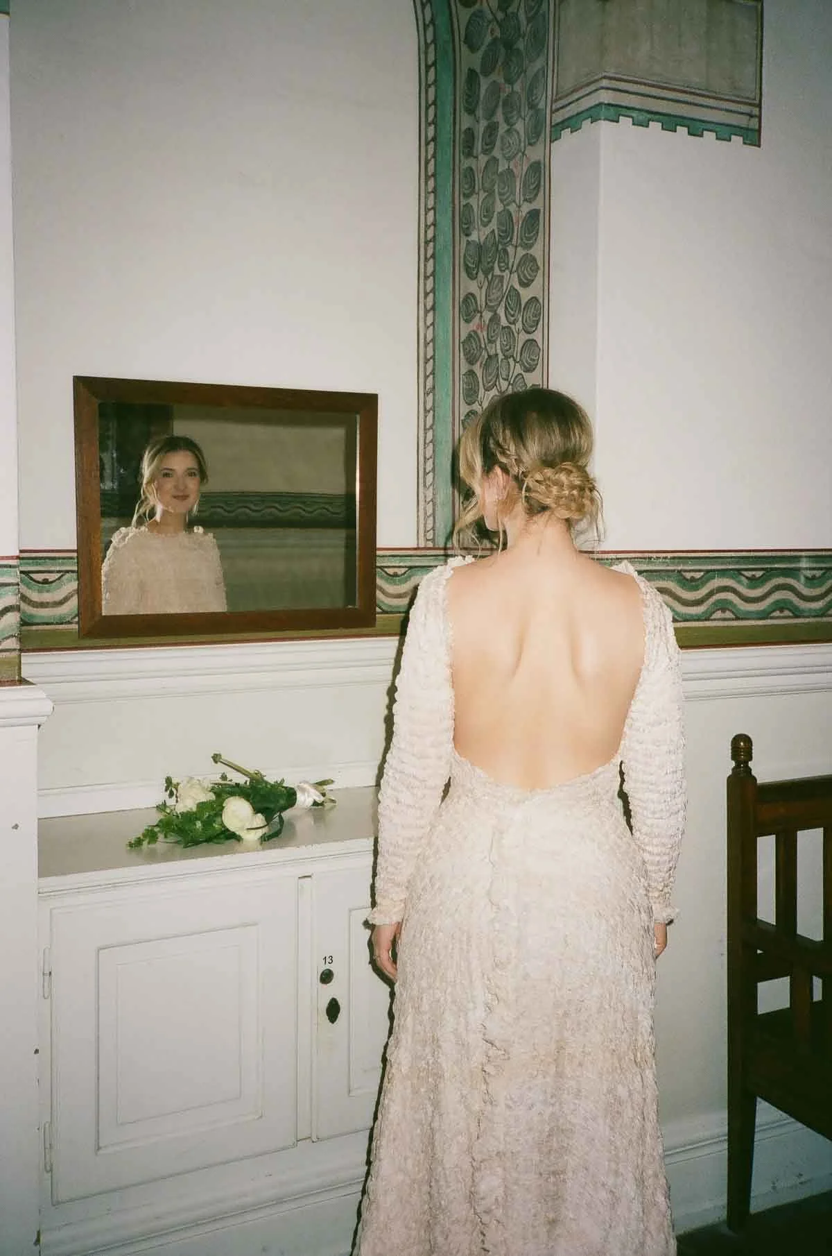 A woman in a cream-colored textured dress with an open back stands in front of a mirror, looking at her reflection.