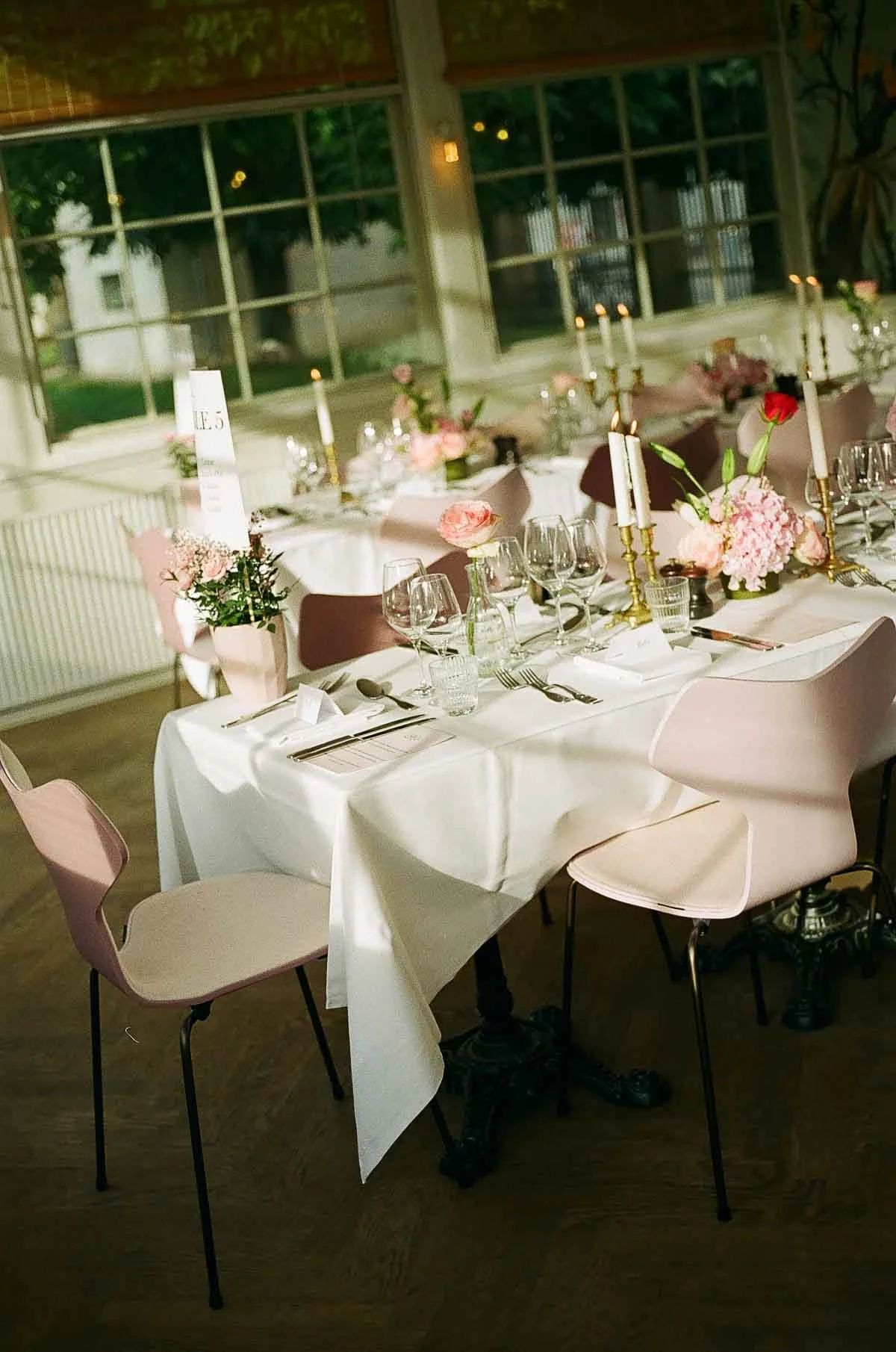 A decorated dining table set for a special occasion features pink and white floral centerpieces, candles, and place settings with wine glasses, cutlery, and napkins in a well-lit room with large windows showing trees outside.