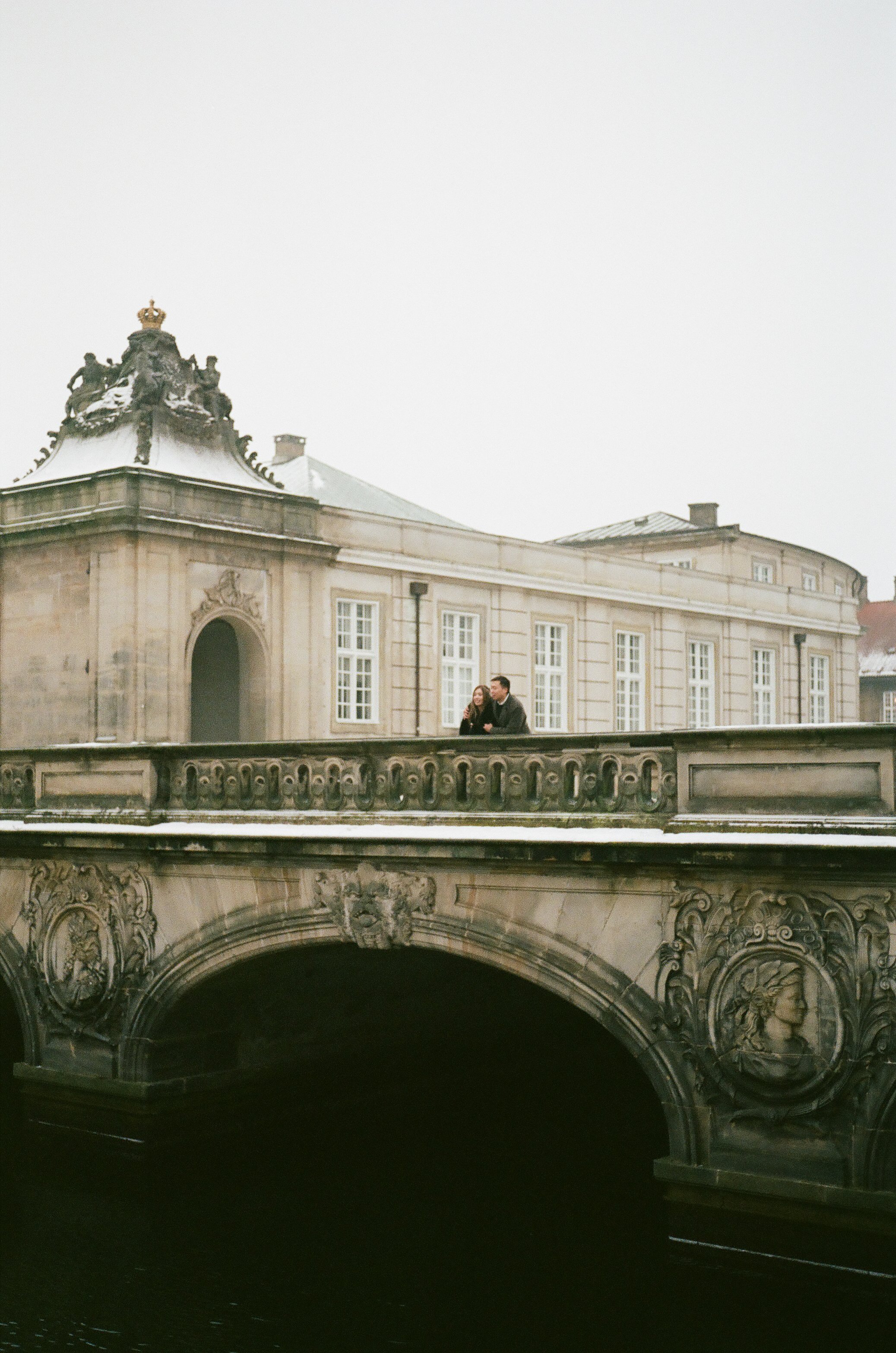 Two people standing on an ornate stone bridge over water in front of a historic building with large windows and decorative stonework, on a foggy day.