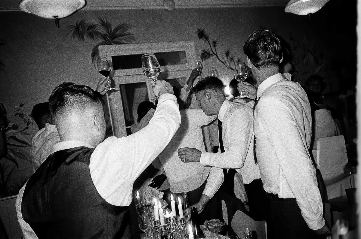 Copenhagen Wedding Photographer. People raising glasses in a toast at a celebratory event, with candles on a table in the foreground.