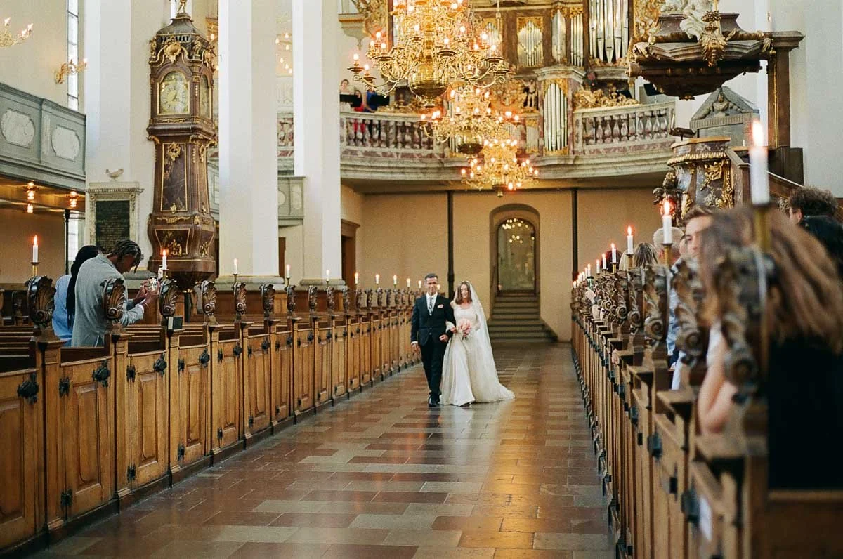 Copenhagen Wedding Photographer. A bride and groom walking down the aisle in a decorated church with chandeliers and wooden pews.