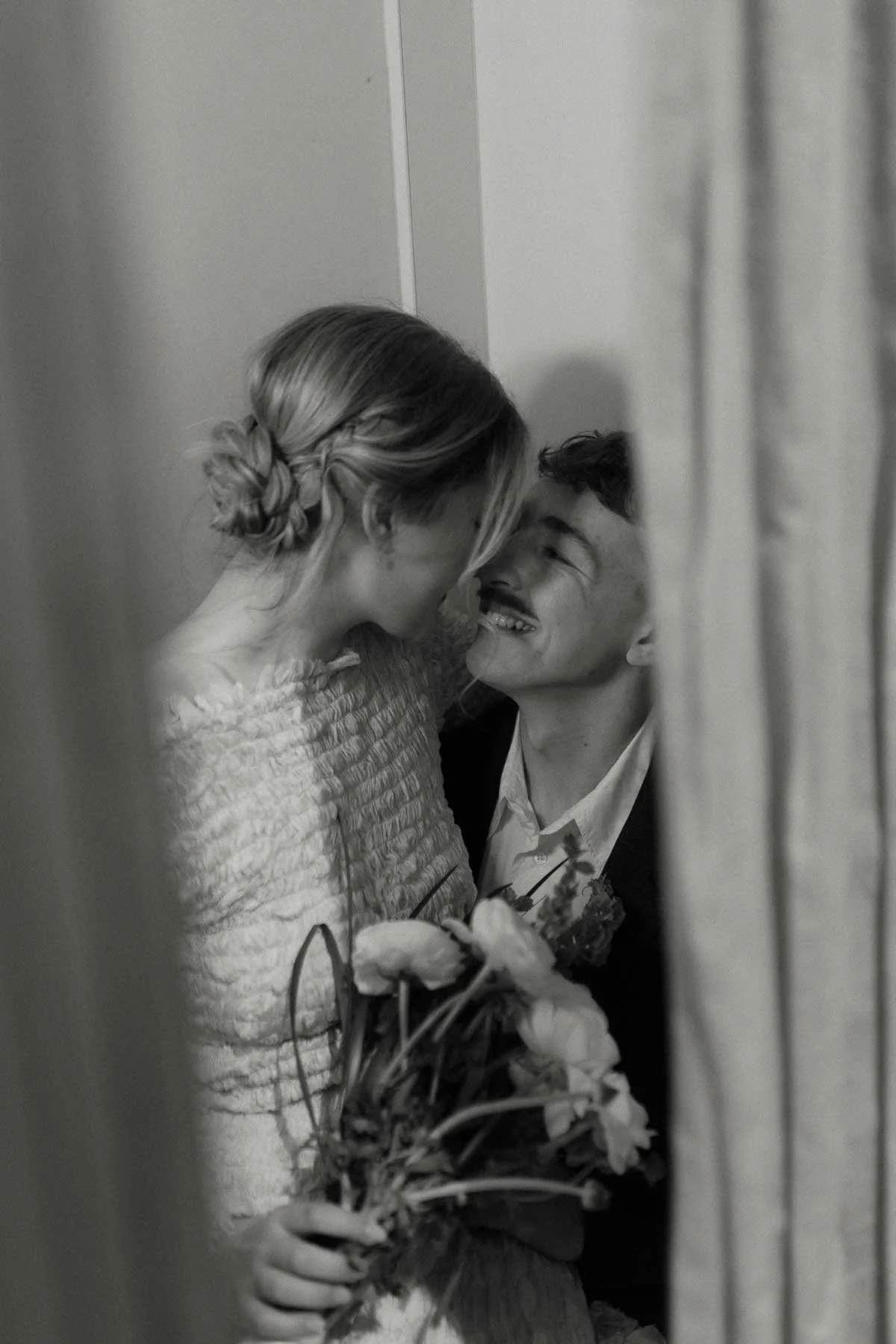 A black and white photo of a couple smiling at each other, close together, with the woman holding a bouquet of flowers.