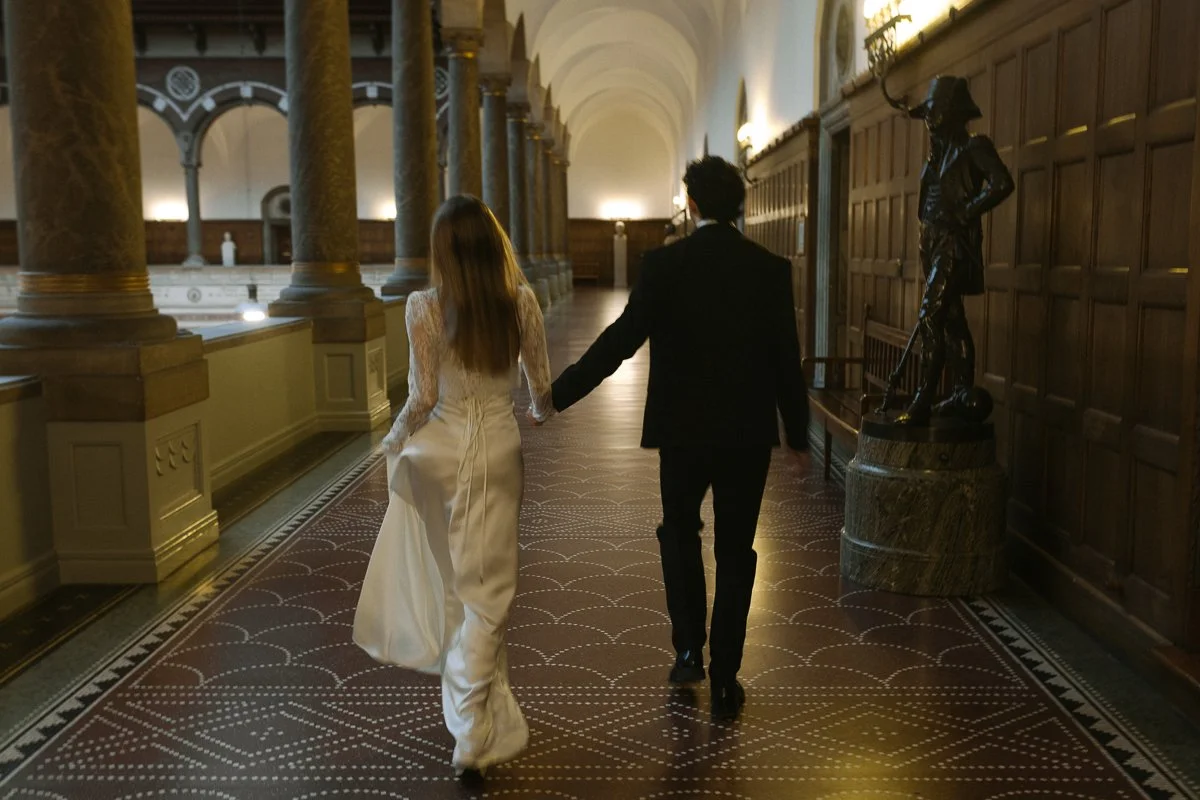 A couple holding hands, walking through a grand hallway with large columns and a statue on a pedestal.