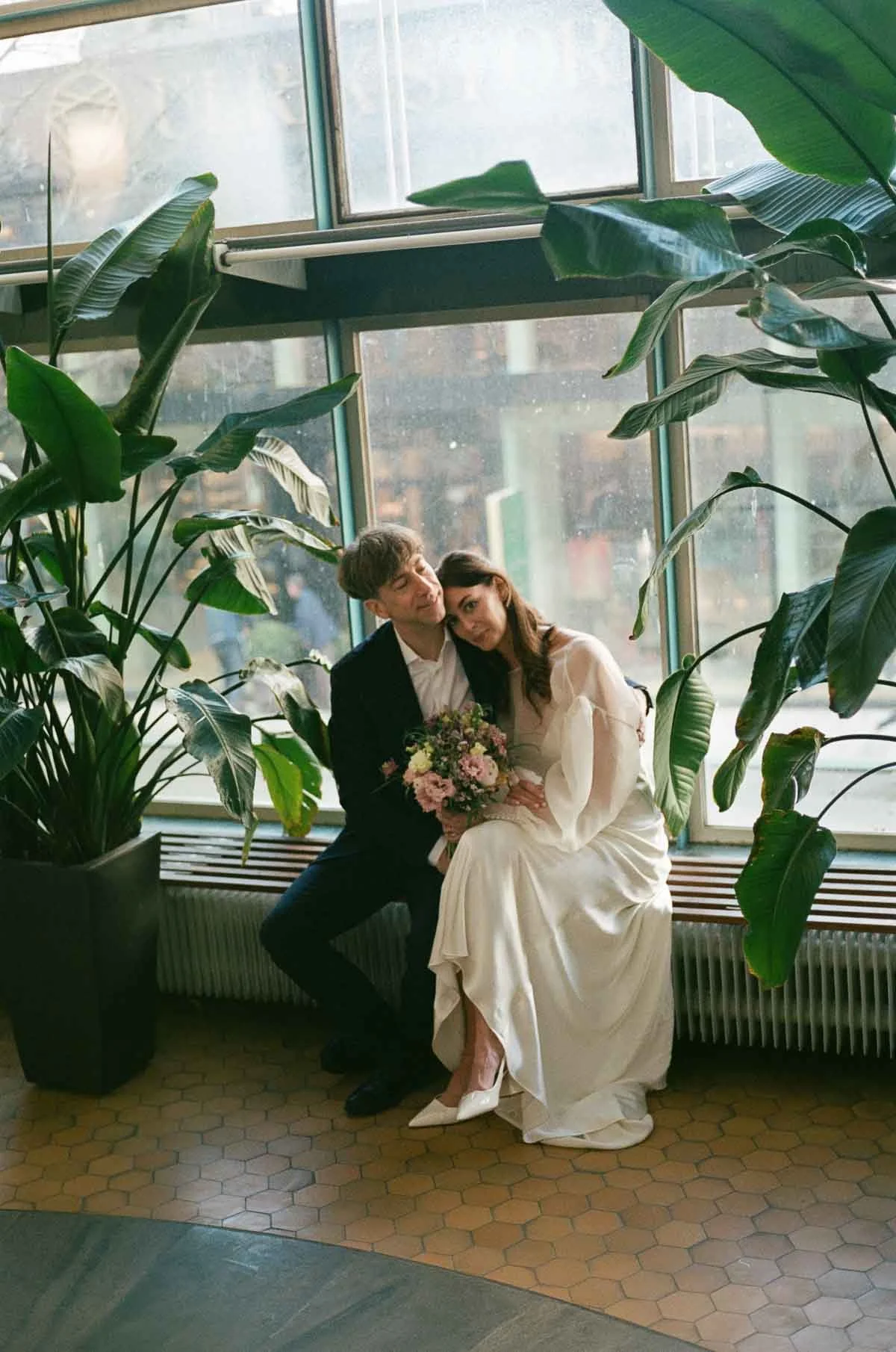 A couple dressed in wedding attire sitting on a bench by a large window, surrounded by large green plants, with the woman holding a bouquet of flowers.
