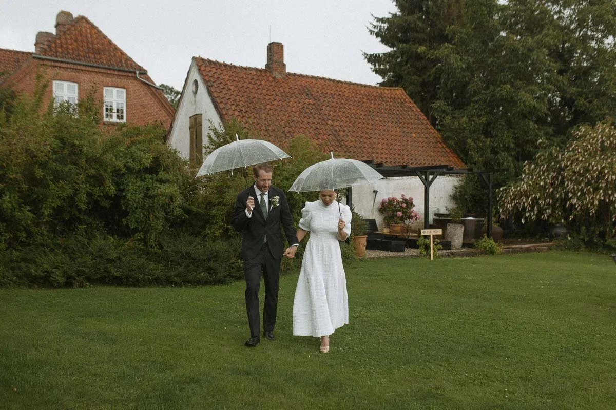 Copenhagen Wedding Photographer. A man in a black suit and a woman in a white dress walking on a grassy lawn with umbrellas, holding hands, in front of a house with a red-tiled roof and surrounding trees on a rainy day.