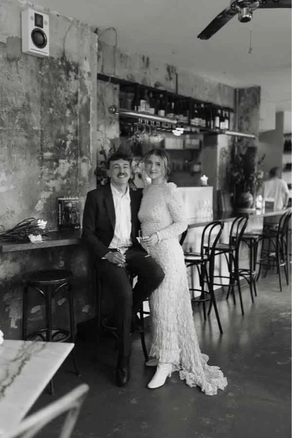 A couple in formal attire at a modern restaurant, with the woman wearing a long, lace dress and the man in a suit, standing close together and smiling.