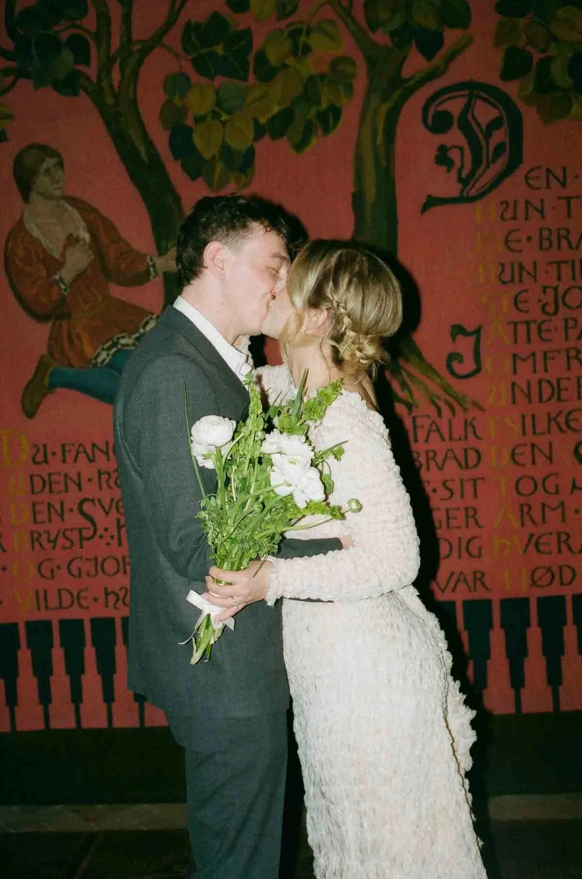 A couple in wedding attire, kissing, with the bride holding a bouquet of white flowers, standing in front of a colorful mural with text.
