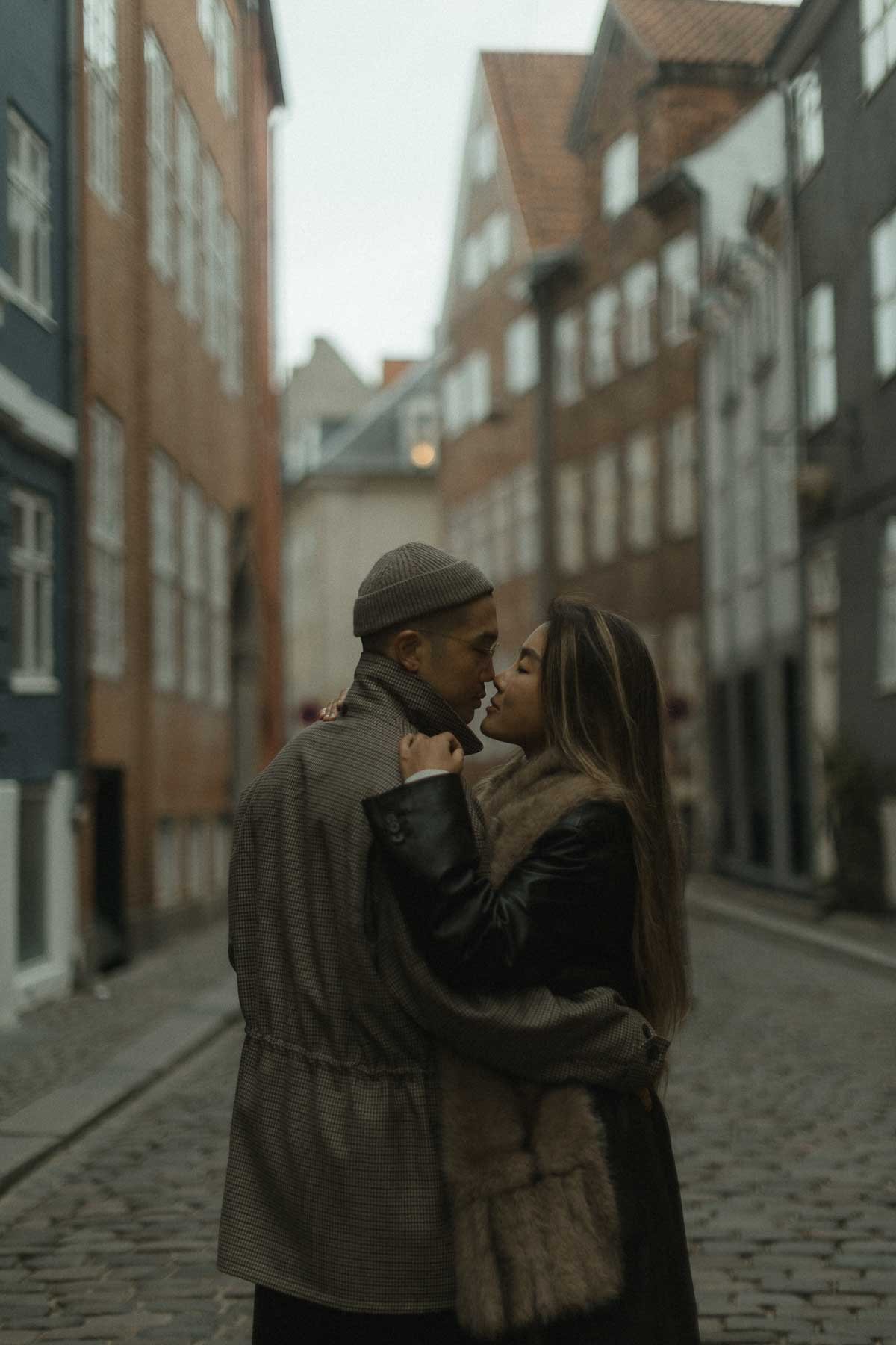 A couple standing close together on a cobblestone street, leaning in for a kiss, surrounded by colorful buildings in the background.