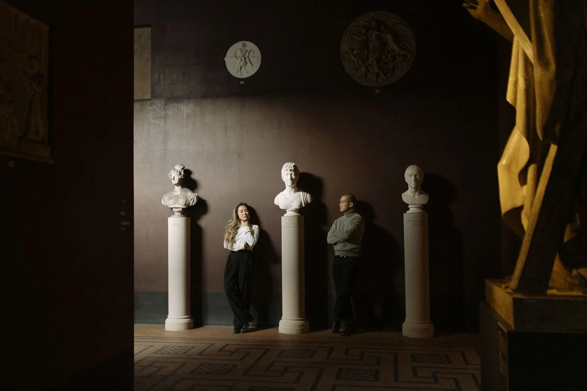 Two people standing in front of classical bust sculptures on pedestals in a museum or gallery with dark walls.