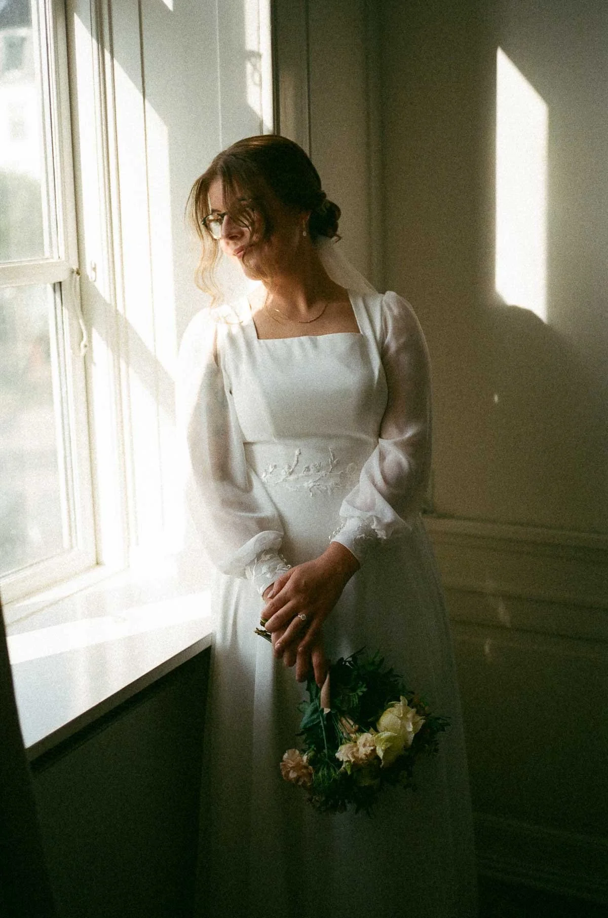 Copenhagen Wedding Photographer. A bride in a white wedding dress standing near a window with sunlight, holding a small bouquet of flowers.