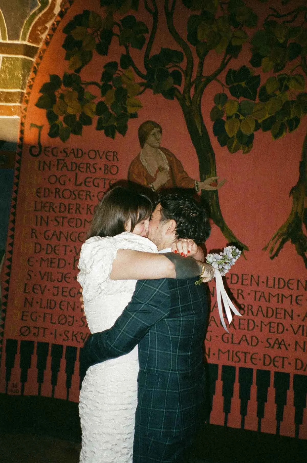 A couple is embracing and about to kiss in front of a mural with a tree and a woman painted on it. The woman in the mural is standing near the tree with her arm extended. The mural features some text in a language other than English, and has a decorative border at the bottom resembling piano keys.
