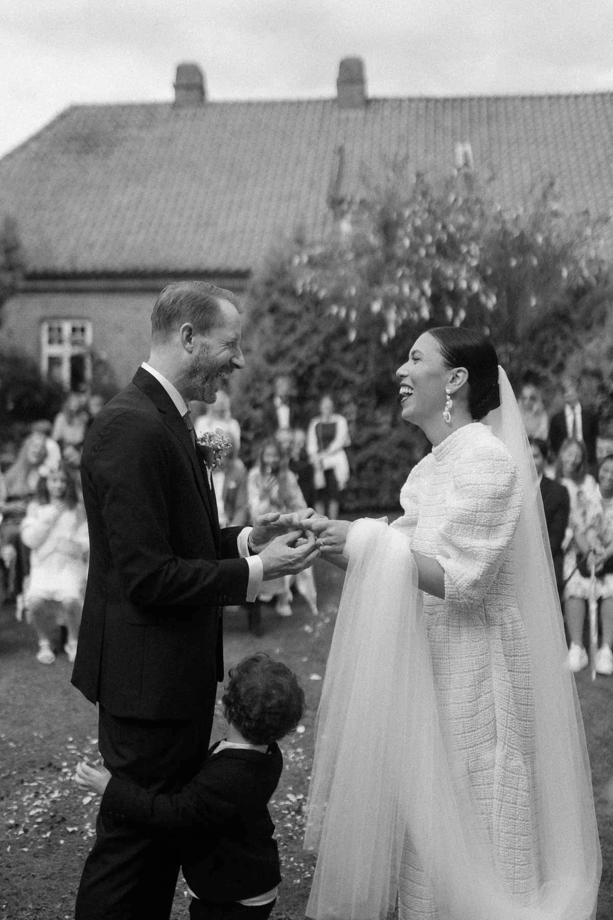 Copenhagen Wedding Photographer. A bride and groom exchanging rings during their outdoor wedding ceremony, with guests in the background.