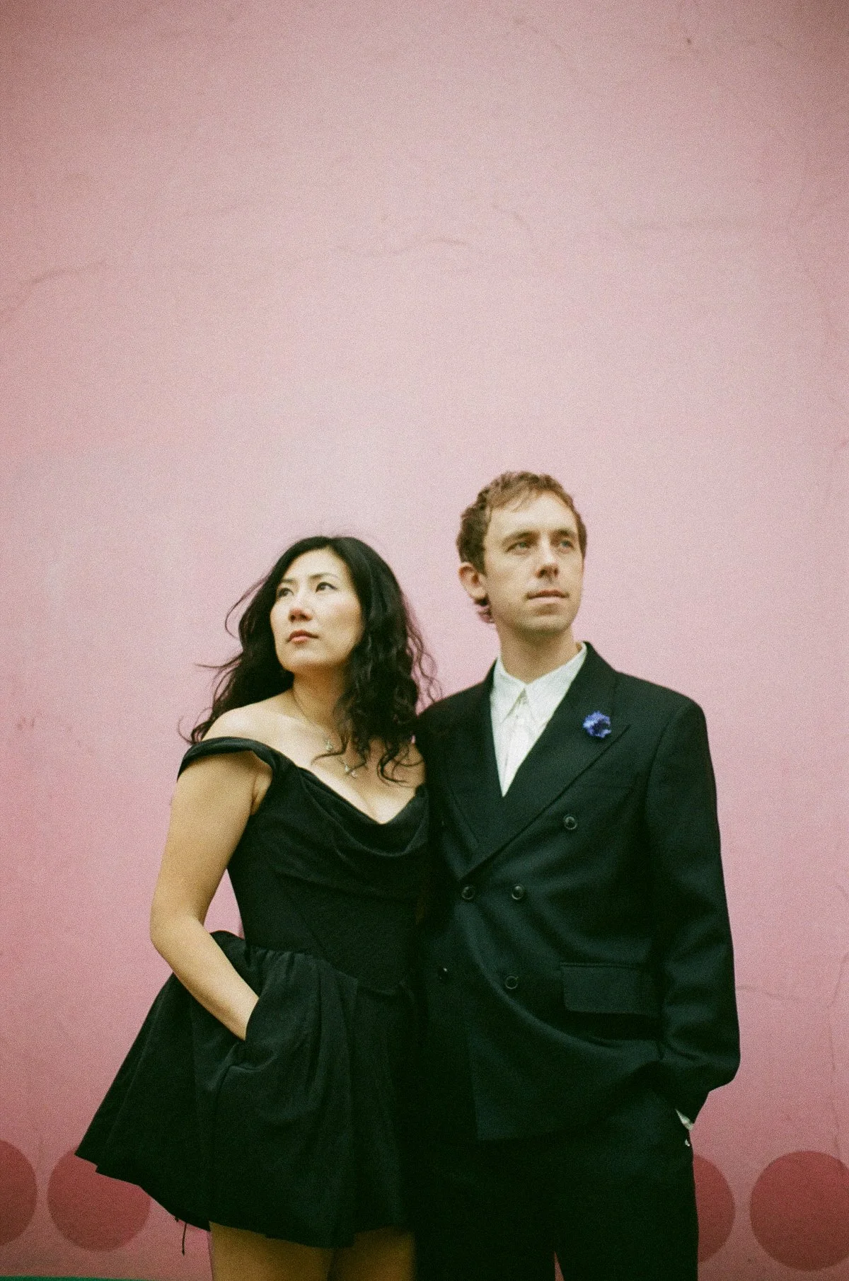 A woman and a man standing in front of a pink wall. The woman is wearing a black dress, and the man is dressed in a black suit with a white shirt, both looking in different directions.