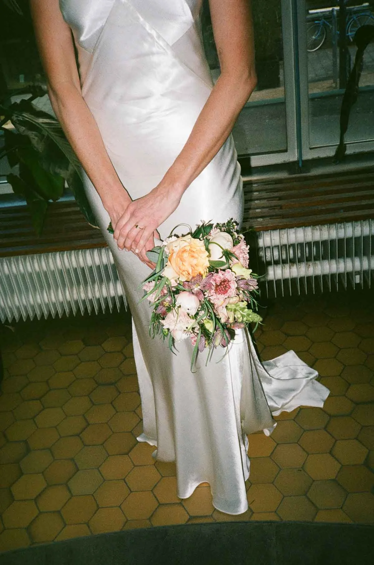 A woman in a shiny white satin dress holding a bouquet of pastel-colored flowers, standing indoors on a hexagonal tile floor near a window.