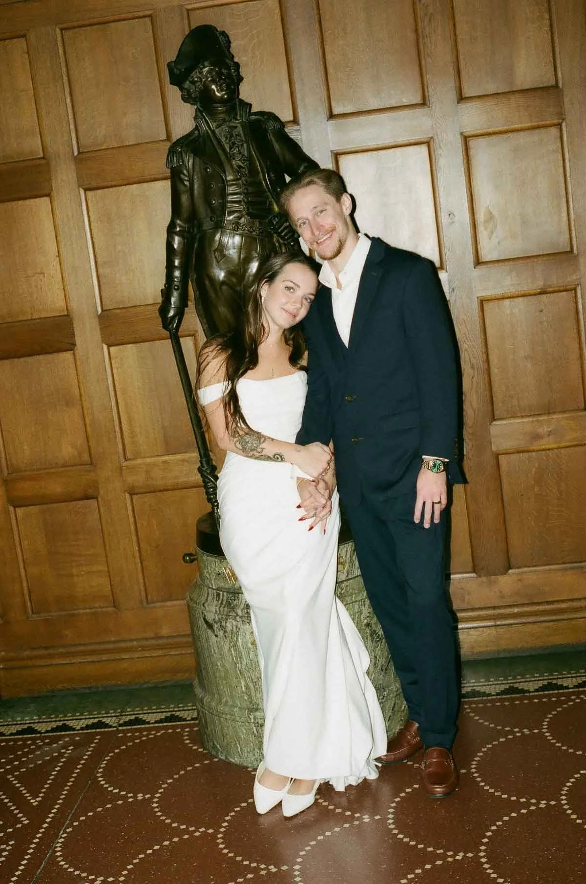 A young woman in a white dress and a young man in a dark suit posing together in front of a Copenhagen Wedding Photographer. wooden wall with a bronze statue of a historic figure, possibly a Revolutionary War soldier, standing behind them.