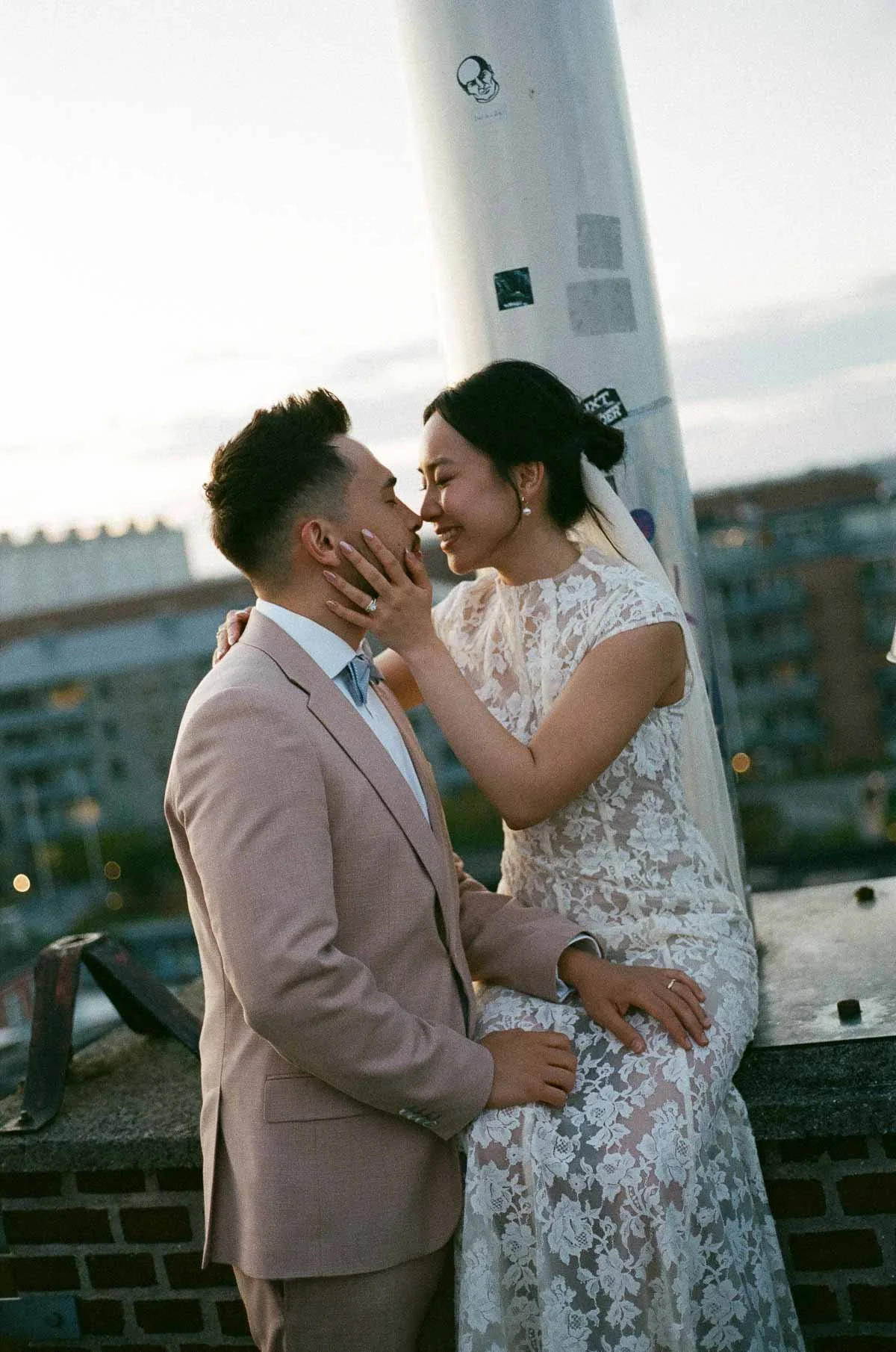 Copenhagen Wedding Photographer. A bride and groom are close together on a rooftop, smiling and touching foreheads, with the bride gently holding the groom's face. The groom is wearing a beige suit with a white shirt and tie, and the bride is in a wh