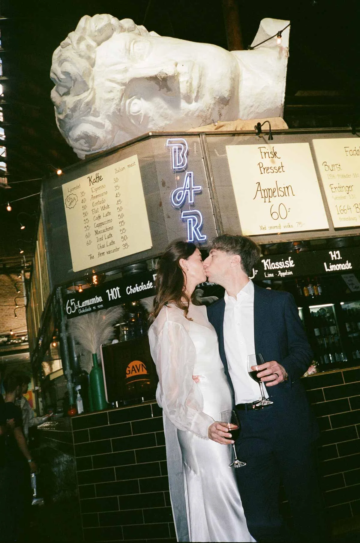 A couple in formal attire kissing inside a bar, with a large white sculpture of a man's face and illuminated 'BAR' sign in the background.