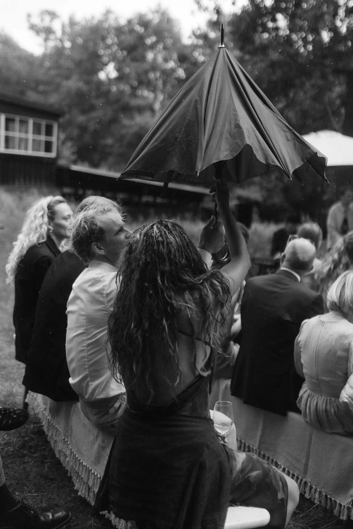 Copenhagen Wedding Photographer. A woman holding an umbrella at an outdoor gathering, with seated people around her, some holding wine glasses, and a building in the background.
